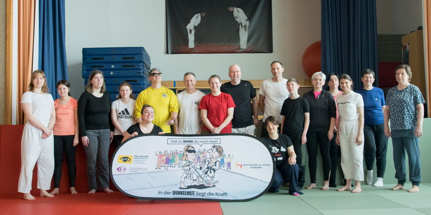 Die buntgemischte Gruppe aus 16 Menschen jeden Alters steht in der Gymnastikhalle der blista vor einem Banner von Pro Retina, DBSV und Ju-Jutsu-Verband. Darauf steht: „In der Dunkelheit liegt die Kraft.“