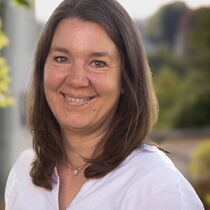 Frau Schneider lächelt freundlich in die Kamera. Sie hat mittellange braune Haare und trägt eine weiße Bluse.