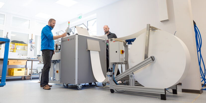 Zwei Mitarbeiter stehen an einem großen Punkschriftdrucker, im Vordergrund ist eine große Papierrolle zu sehen.