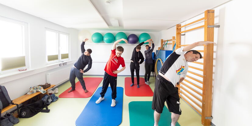 Vier Menschen unterschiedlichen Alters dehnen sich angeleitet durch die Motologin in einem Gymnastikraum, die Trainerin korrigiert.