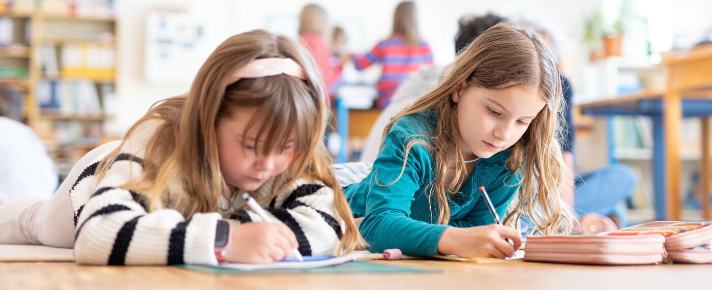 Zwei Schülerinnen liegen auf dem Fußboden und schreiben etwas.