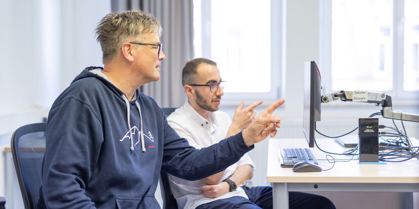 Ein Ausbilder und ein Azubi sitzen vor einem Bildschirm in einem Unterrichtsraum und zeigen auf den Bildschirminhalt.