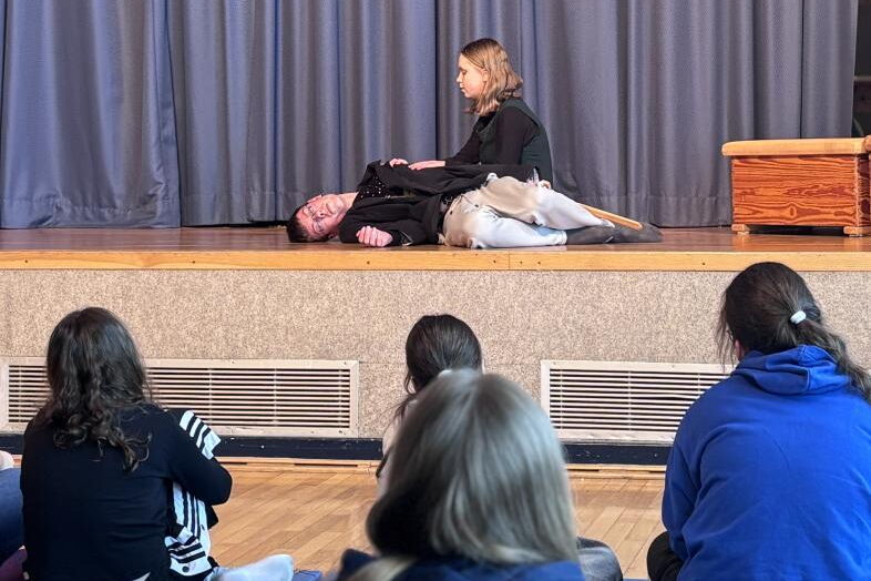 Auf einer Bühne sind zwei Schüler*innen ins Theaterspiel vertieft, einer liegt reglos am Boden, während die andere Person neben ihm kniet.