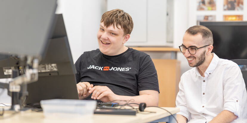 Zwei junge Männer sitzen gemeinsam an einem Pc und lächeln.
