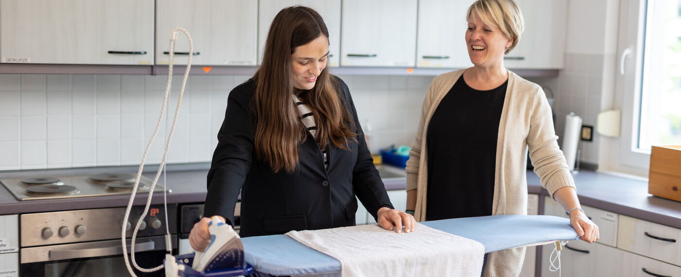 Eine Teilnehmerin steht an einem Bügelbrett und greift nach einem Bügeleisen, während eine Lehrkraft neben ihr steht.