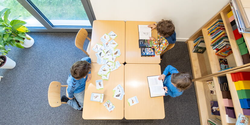 In einem Arbeitsraum blickt man von oben auf einen Tisch, an dem drei Kinder sitzen und mit unterschiedlichen Materialien arbeiten.
