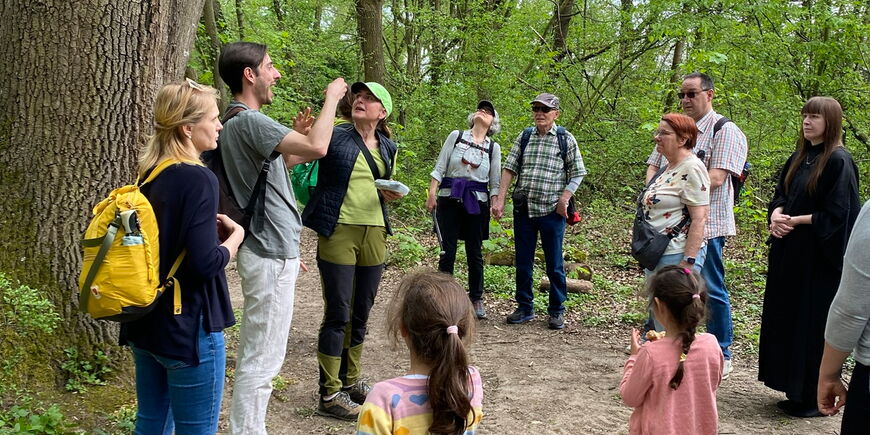 Eine Gruppe von Erwachsenen und zwei Kindern steht auf einem Waldweg unter einem großen Baum. Eine Person in der Mitte scheint der Gruppe etwas zu erklären.