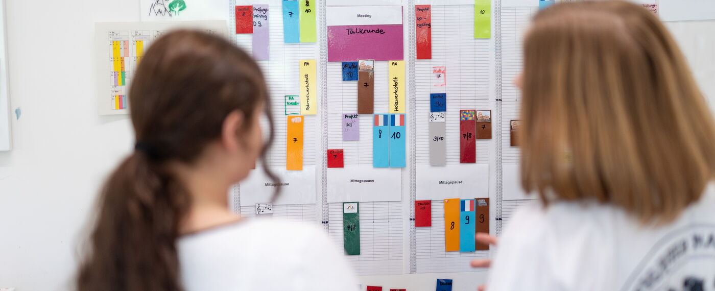 Zwei Schülerinnen stehen vor einem großen Wochenplan mit bunten Zetteln.