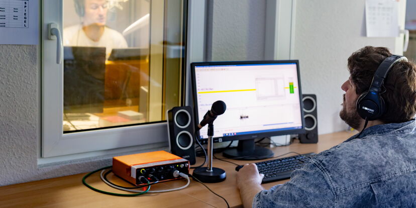 Ein Hörbuchsprecher hinter einem Fenster einer Sprecherkabine, ein Kollege mit Headset außerhalb spricht mit ihm.