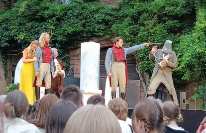 5 historisch gekleidete Schauspieler*innen. Einer der Schauspieler hält einem anderen eine Waffe an den Kopf, welcher wiederum einen Eimer auf dem Kopf hat und eine Katze im Arm trägt.