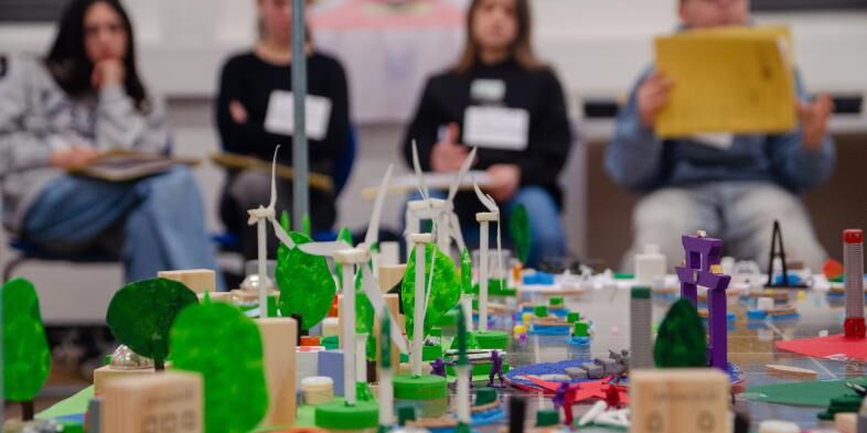 Im Vordergrund sieht man das Board mit diversen bunten Spielfiguren, im Hintergrund sitzen Schüler*innen.