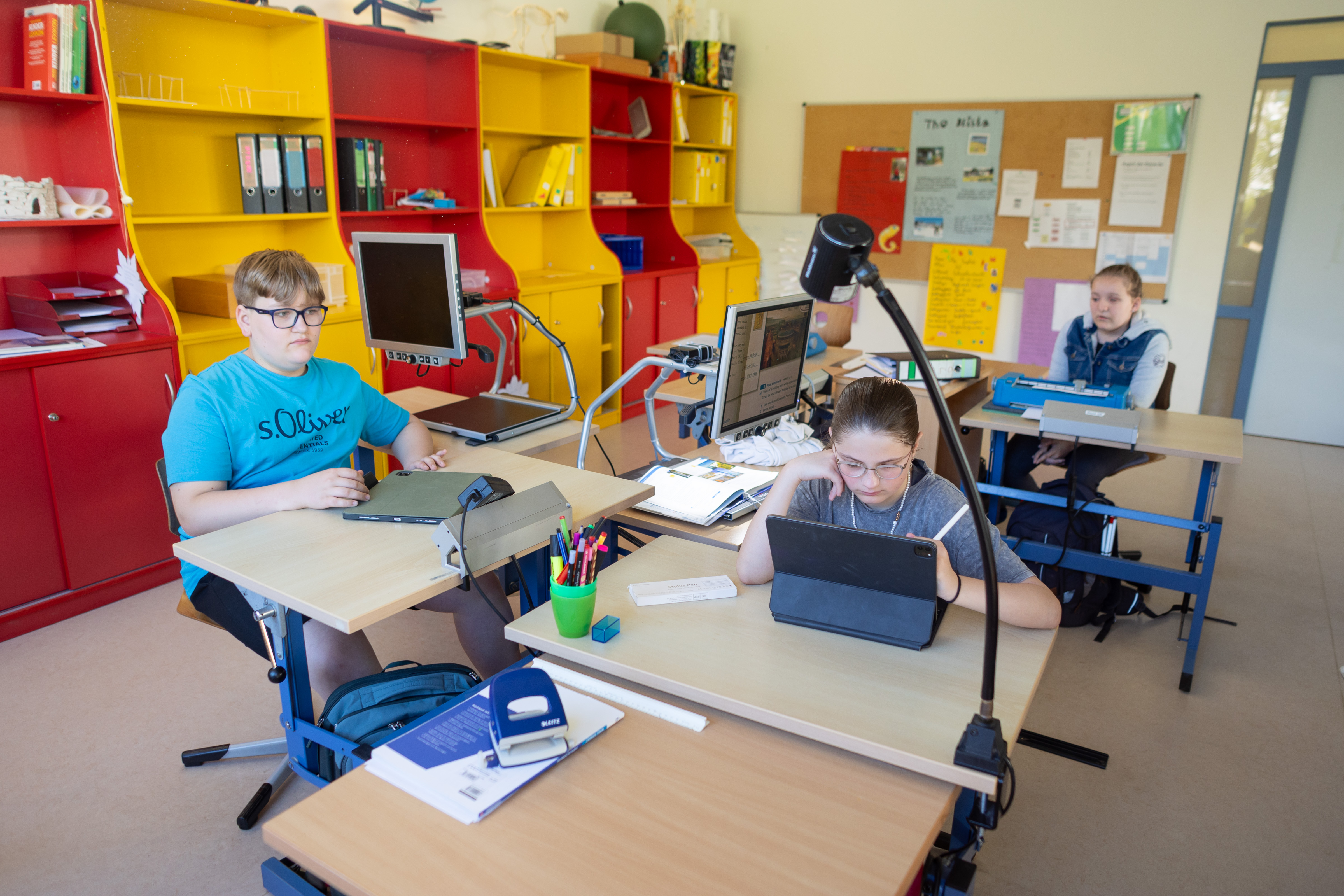 Drei Schüler*innen sitzen an ihren Arbeitsplätzen und arbeiten mit unterschiedlichen Medien.