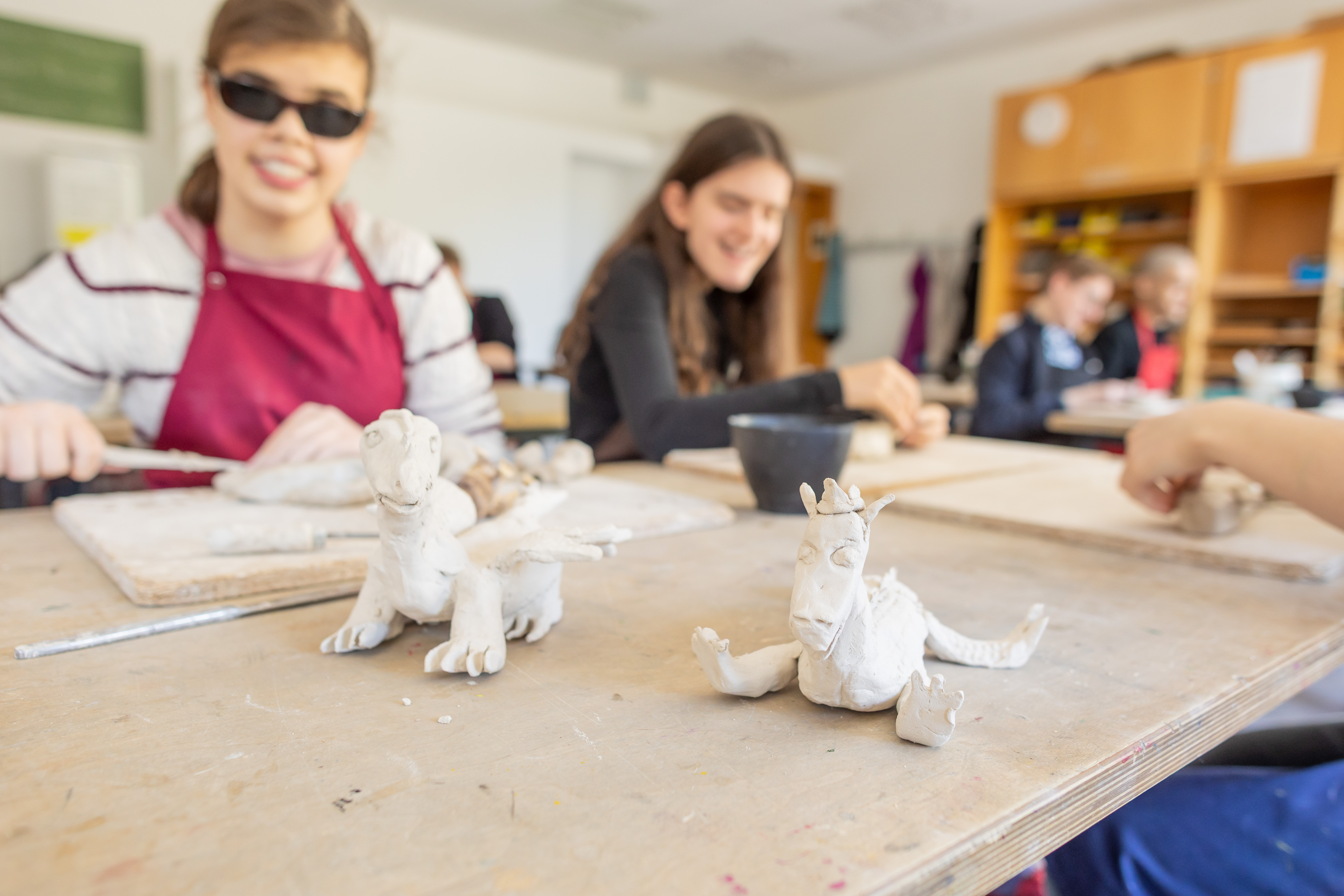 Im Vordergrund stehen zwei getöpferte Drachen, im Hintergrund arbeiten Schüler*innen an Ton.