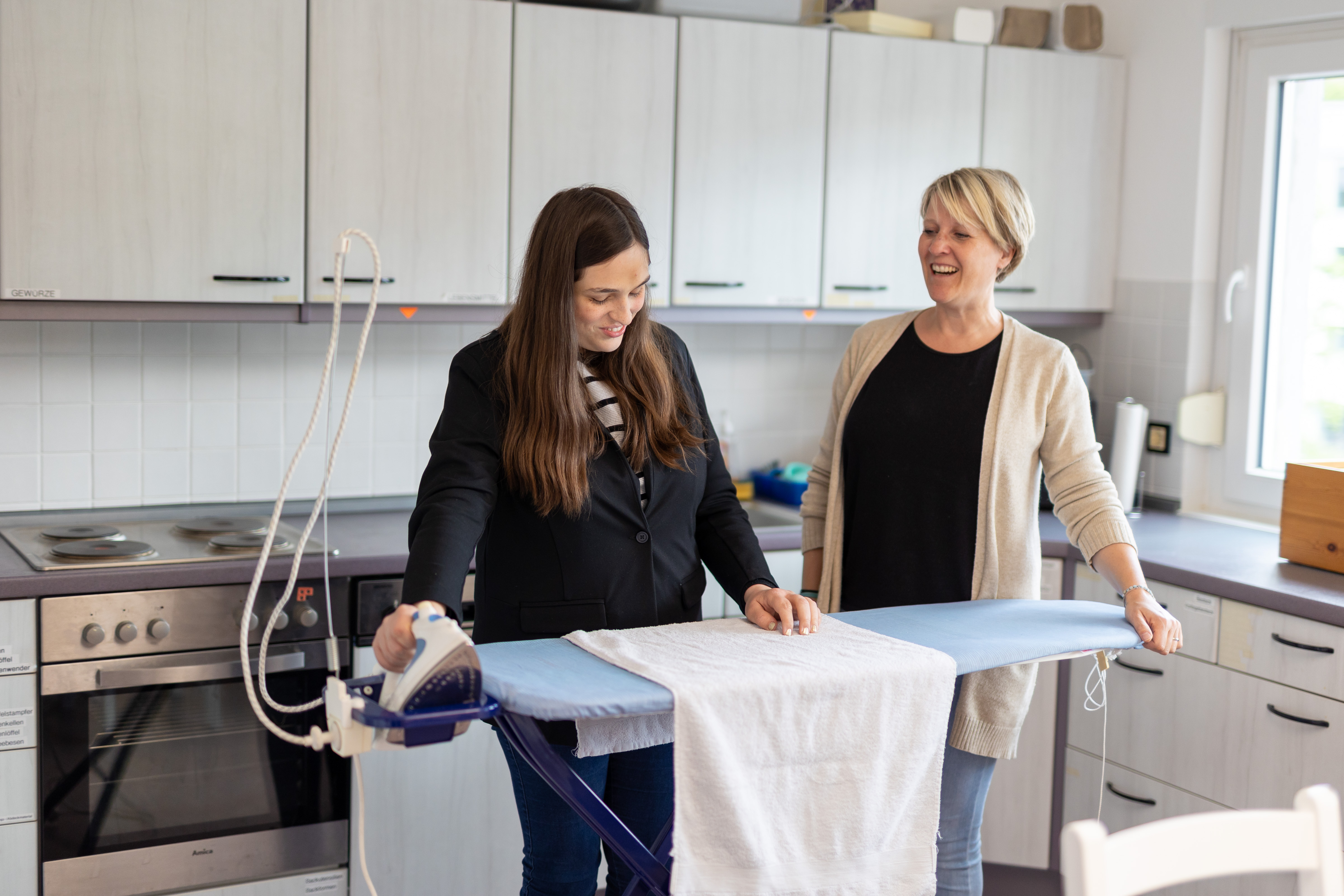 Eine Teilnehmerin steht an einem Bügelbrett und greift nach einem Bügeleisen, während eine Lehrkraft neben ihr steht.